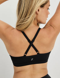 A woman shown from the back wearing a black sports bra with crisscross shoulder straps and a small silver logo at the center back, against a light gray studio background.